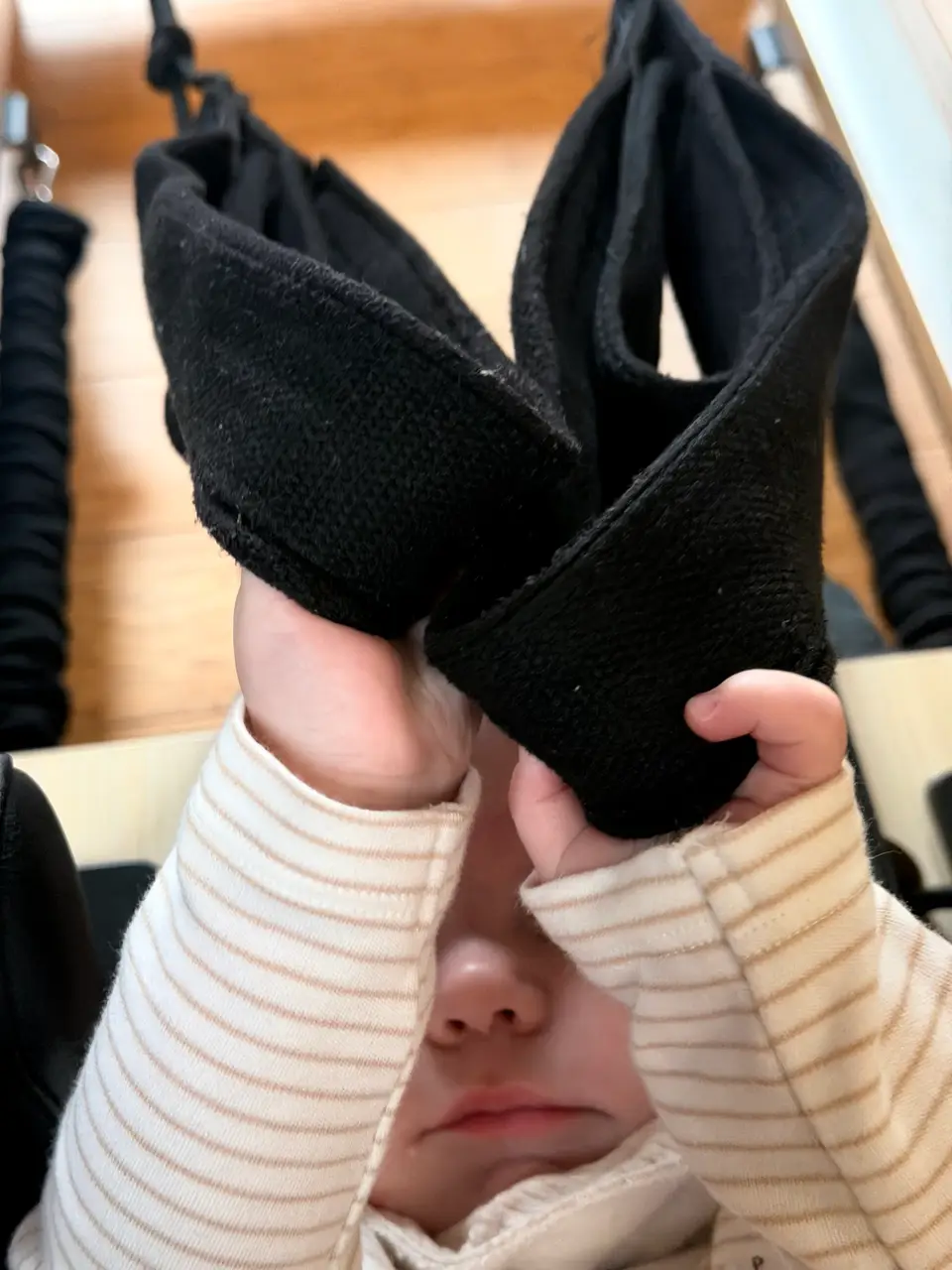 A baby in a striped babygrow holding the reformer's foot straps up to their face