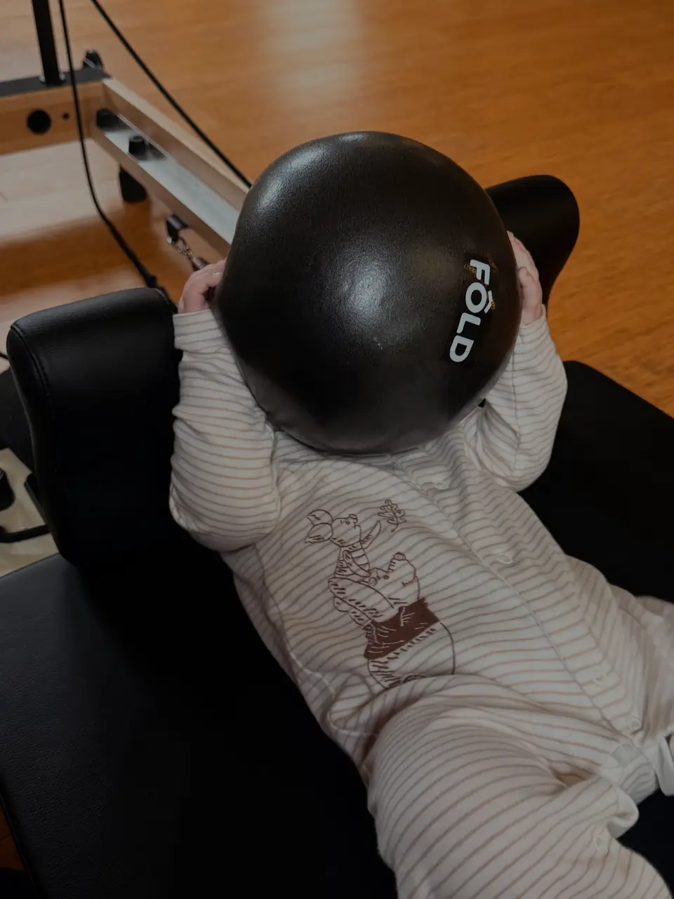 A baby on a reformer, hugging a black exercise ball