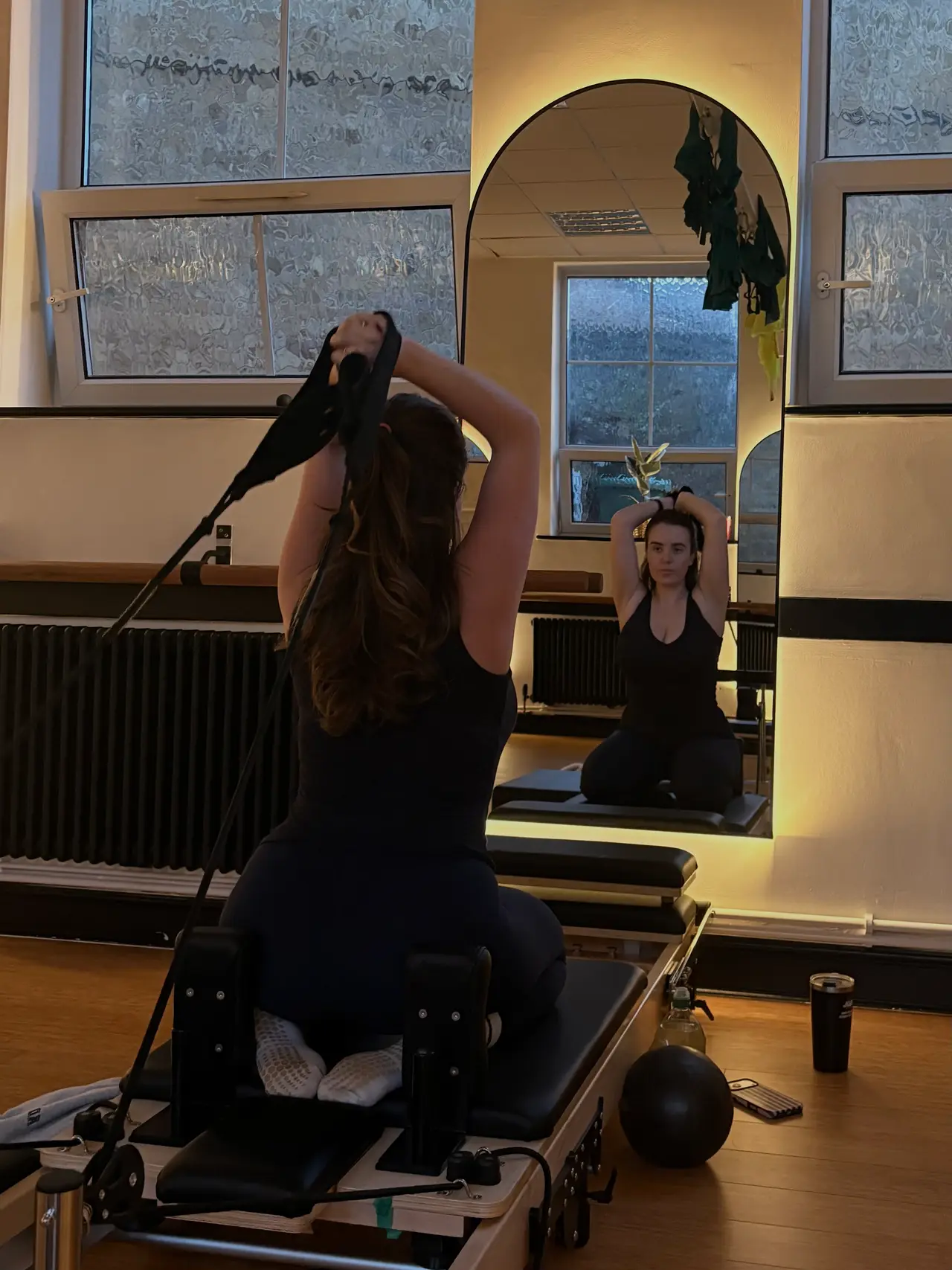 A member kneeling on a reformer, pulling the straps overhead, facing the studio mirror